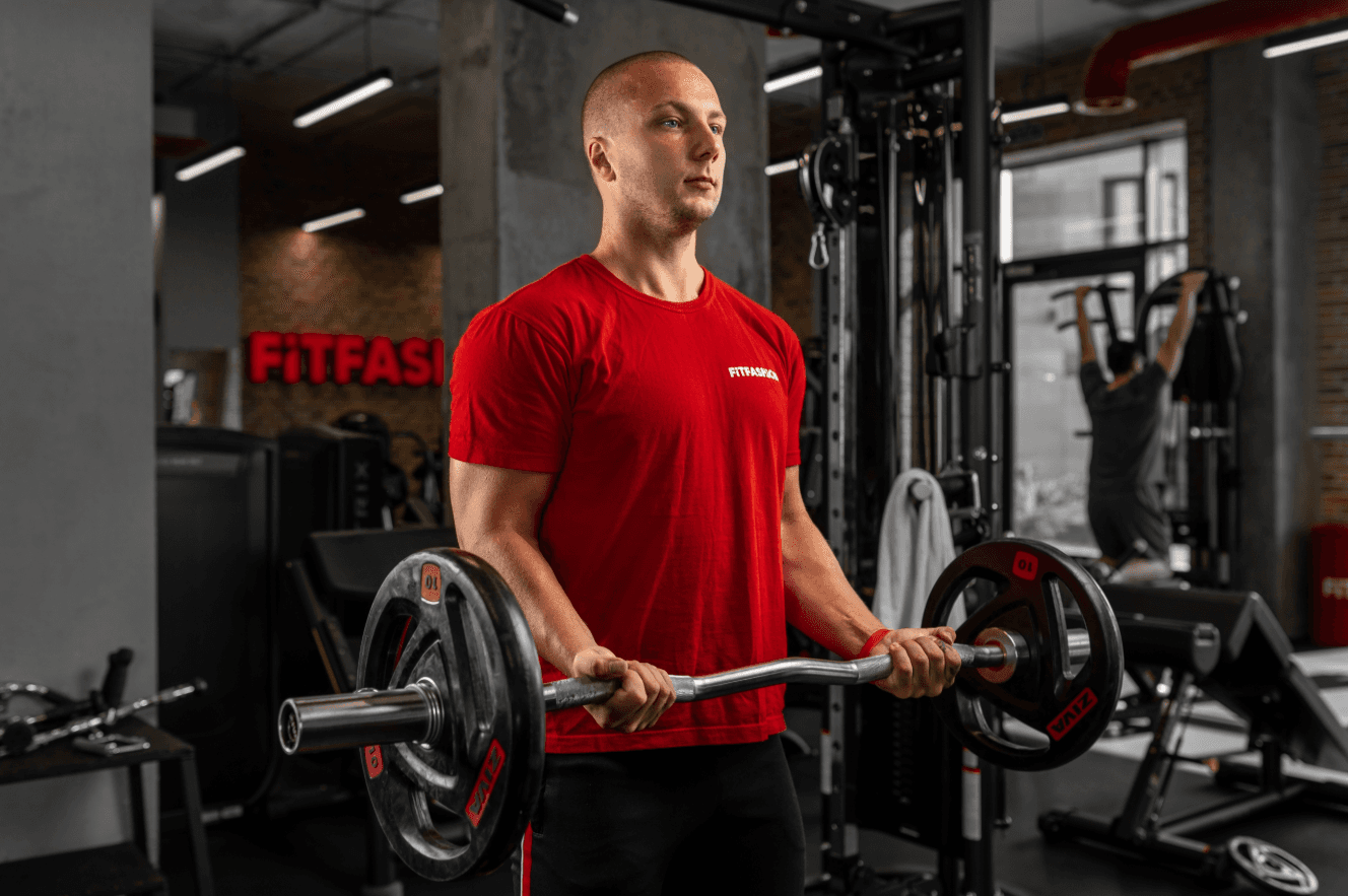 A man in a red shirt performs a barbell curl in a gym.