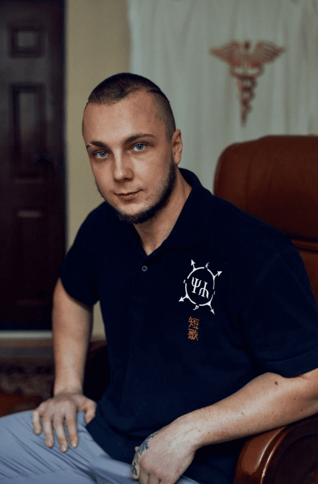 Young man in a dark shirt seated in an office with a medical symbol behind him.