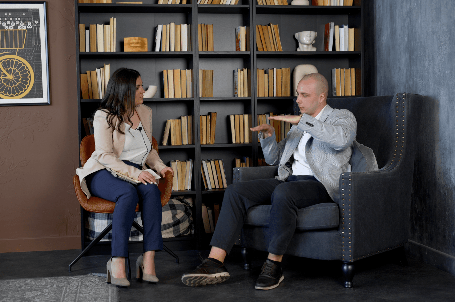 Man and woman seated in armchairs during an interview in front of bookshelves.