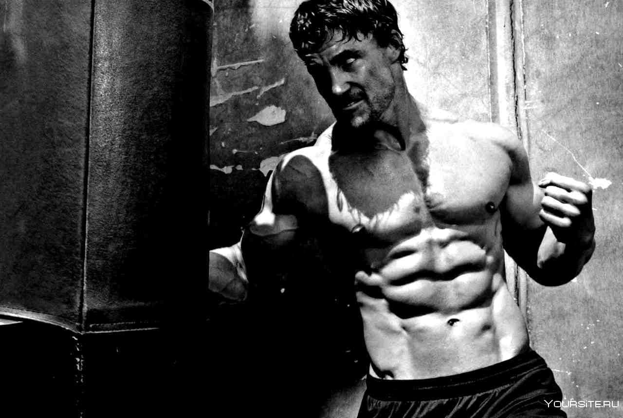 A black-and-white photograph of a muscular shirtless man in a worn industrial interior.