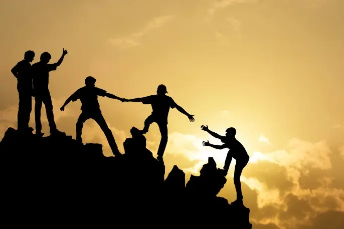 Five silhouetted people help one another climb a rocky slope at sunset
